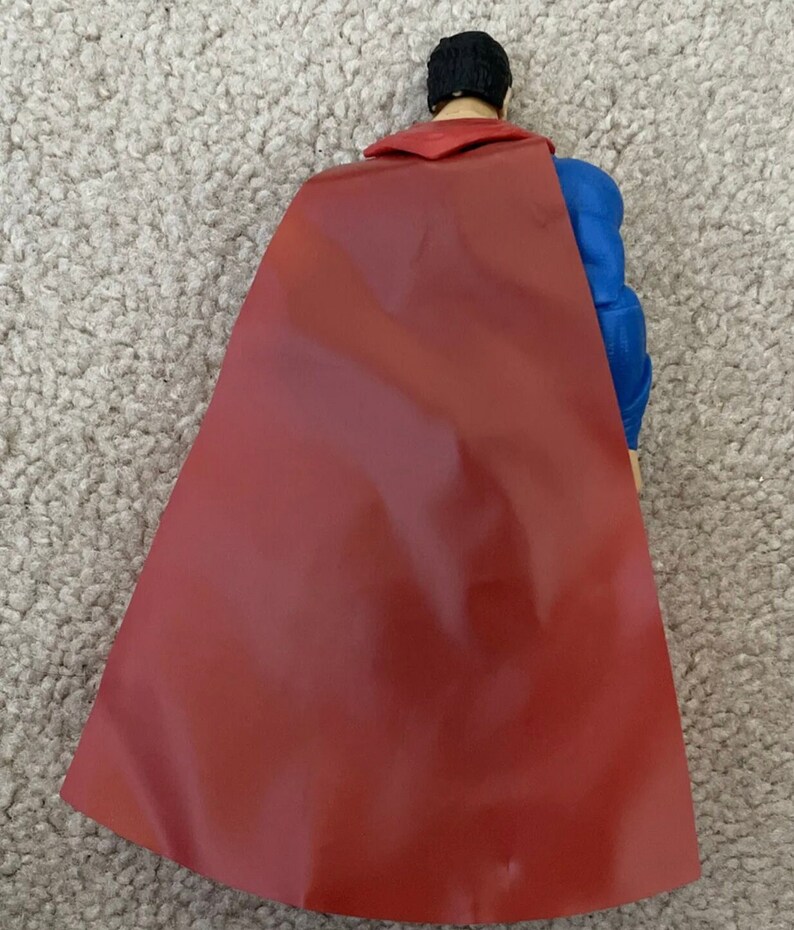 May include: A Superman action figure viewed from the back, showcasing a red cape and blue suit. The figure has black hair and a red collar. The cape is a deep red color.