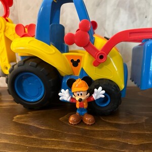 May include: A yellow, blue, and red toy construction vehicle with black wheels. A Mickey Mouse figurine in an orange hard hat and work clothes stands in front of the vehicle. The vehicle has a Mickey Mouse logo.