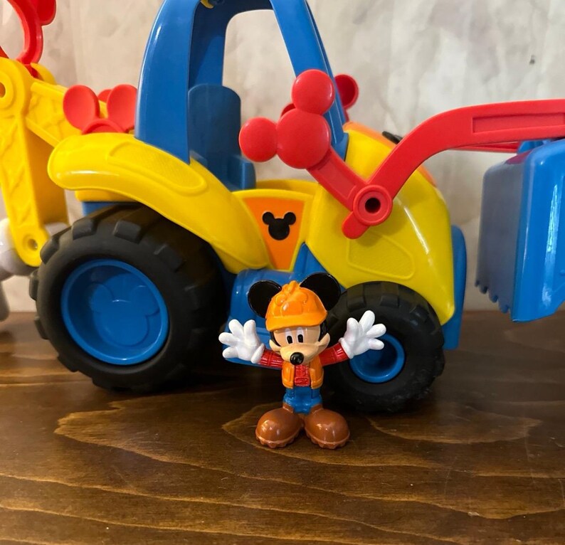 May include: A yellow, blue, and red toy construction vehicle with black wheels. A Mickey Mouse figurine in an orange hard hat and work clothes stands in front of the vehicle. The vehicle has a Mickey Mouse logo.