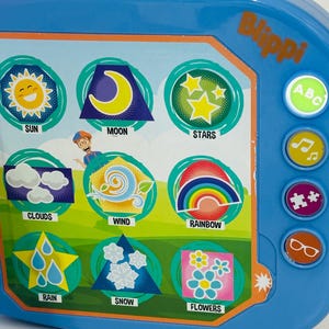 May include: A blue Blippi-themed educational toy with colorful illustrations of the sun, moon, stars, clouds, wind, rainbow, rain, snow, and flowers. The toy has buttons with ABC, music notes, puzzle pieces, and glasses icons.