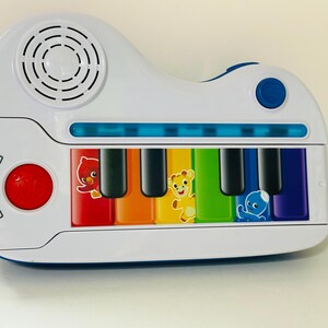Pode incluir: Um piano de brinquedo branco e azul com teclas coloridas e gr&aacute;ficos de animais. O piano tem um bot&atilde;o vermelho, um alto-falante e o texto "Baby Einstein". As teclas s&atilde;o vermelhas, laranja, amarelas, verdes, azuis e roxas.