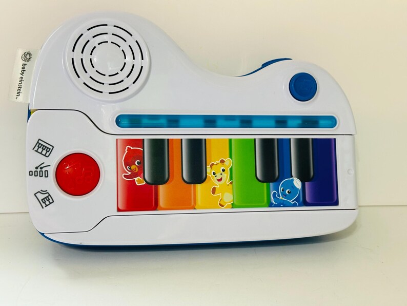 Pode incluir: Um piano de brinquedo branco e azul com teclas coloridas e gr&aacute;ficos de animais. O piano tem um bot&atilde;o vermelho, um alto-falante e o texto "Baby Einstein". As teclas s&atilde;o vermelhas, laranja, amarelas, verdes, azuis e roxas.