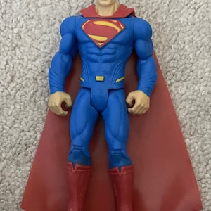 May include: Superman action figure in a classic blue suit with a red cape and boots. The figure has a yellow and red Superman logo on the chest. The figure is approximately 6 inches tall.