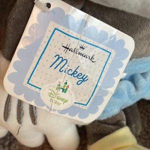 May include: A plush Mickey Mouse toy with a Hallmark and Disney Baby tag. The tag is light blue with the name "Mickey" in script. The toy has gray, white, and yellow accents.
