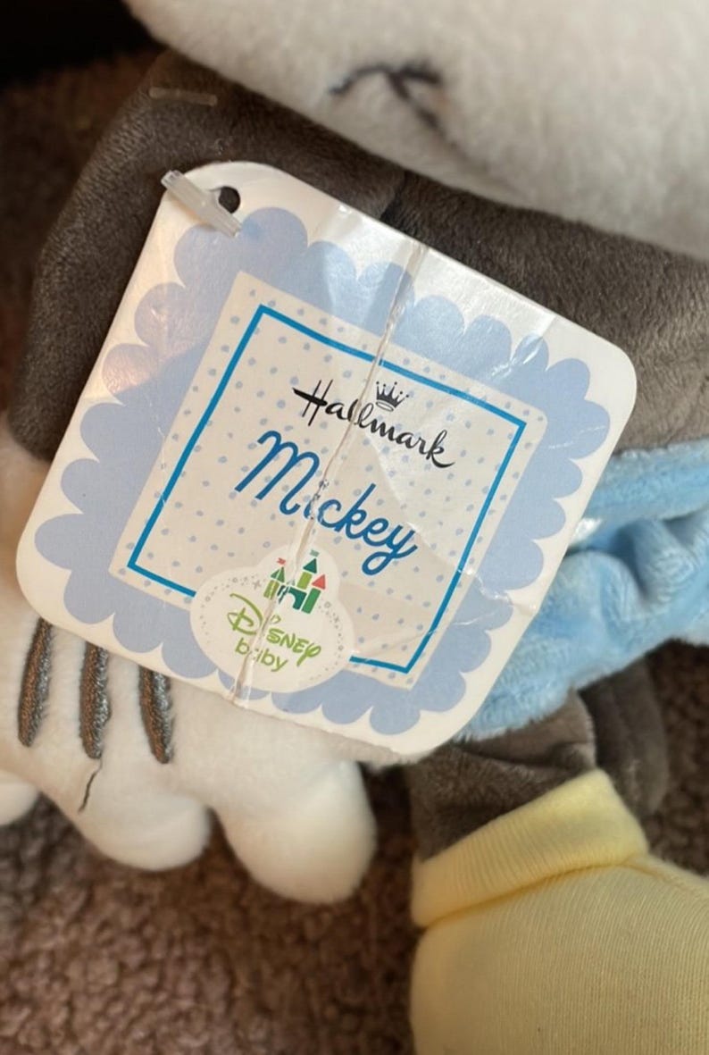 May include: A plush Mickey Mouse toy with a Hallmark and Disney Baby tag. The tag is light blue with the name "Mickey" in script. The toy has gray, white, and yellow accents.