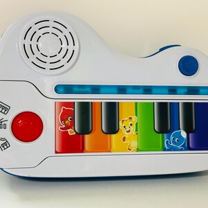 Pode incluir: Um piano de brinquedo Baby Einstein branco e azul com teclas coloridas e ilustra&ccedil;&otilde;es de animais. O brinquedo possui um alto-falante, um bot&atilde;o vermelho e um bot&atilde;o azul. As teclas s&atilde;o em cores do arco-&iacute;ris.