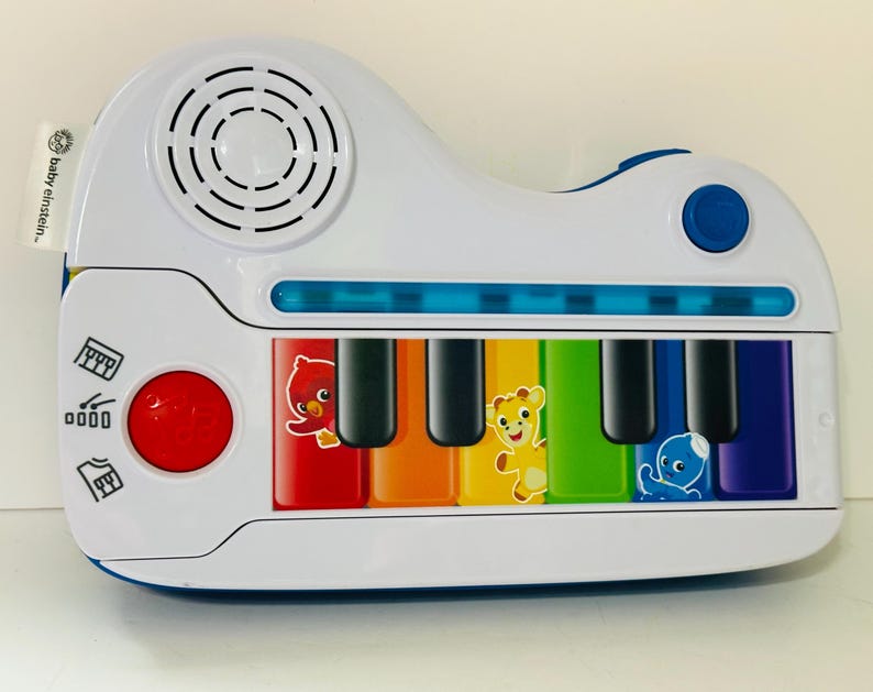 Pode incluir: Um piano de brinquedo Baby Einstein branco e azul com teclas coloridas e ilustra&ccedil;&otilde;es de animais. O brinquedo possui um alto-falante, um bot&atilde;o vermelho e um bot&atilde;o azul. As teclas s&atilde;o em cores do arco-&iacute;ris.