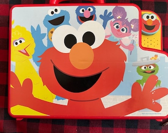 Puede incluir: Maleta de juguete roja de Barrio Sésamo con Elmo, Big Bird, Cookie Monster y otros personajes. La maleta tiene un asa roja y una colorida ilustración de los personajes de Barrio Sésamo sobre un fondo azul claro.