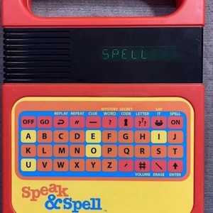 May include: A vintage red Speak & Spell electronic learning toy. The toy features a keyboard with alphabet letters, function buttons, and a digital display showing the word "SPELL". The Speak & Spell logo is visible on the front.