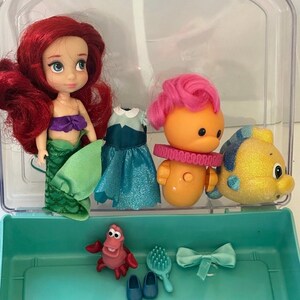May include: Ariel doll with red hair, a purple top, and green tail. Includes a Flounder figure, a Sebastian figure, a dress, shoes, a hairbrush, and a bow. All items are inside a clear plastic case with a teal base.
