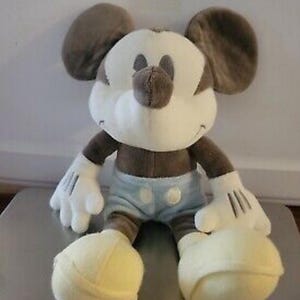 May include: A plush Mickey Mouse doll in a seated position. The doll features a white face, hands, and feet, with a brown nose, ears, and body. It wears light blue shorts with white buttons. The doll is a classic Disney character.