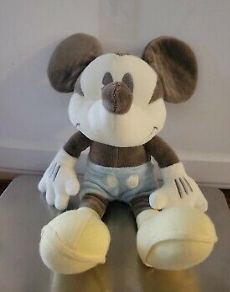 May include: A plush Mickey Mouse doll in a seated position. The doll features a white face, hands, and feet, with a brown nose, ears, and body. It wears light blue shorts with white buttons. The doll is a classic Disney character.