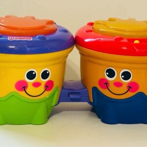 May include: A pair of colorful Fisher-Price toy bongos. The drums are yellow with blue, orange, and red accents. Each drum has a smiling face with pink cheeks and black eyes. The drums are connected by a purple plastic piece.