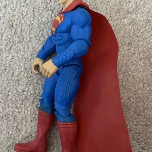 May include: A Superman action figure in a classic blue suit with a red cape and boots. The figure has a detailed face and the iconic Superman logo on the chest. The figure is posed in a dynamic stance.