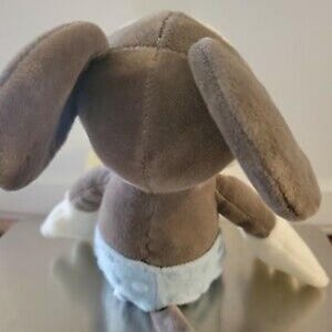 May include: A plush toy bunny, viewed from the back, featuring long, floppy ears and a brown body. The bunny wears a light blue diaper with white trim and has white paws. The toy is made of soft, plush material.