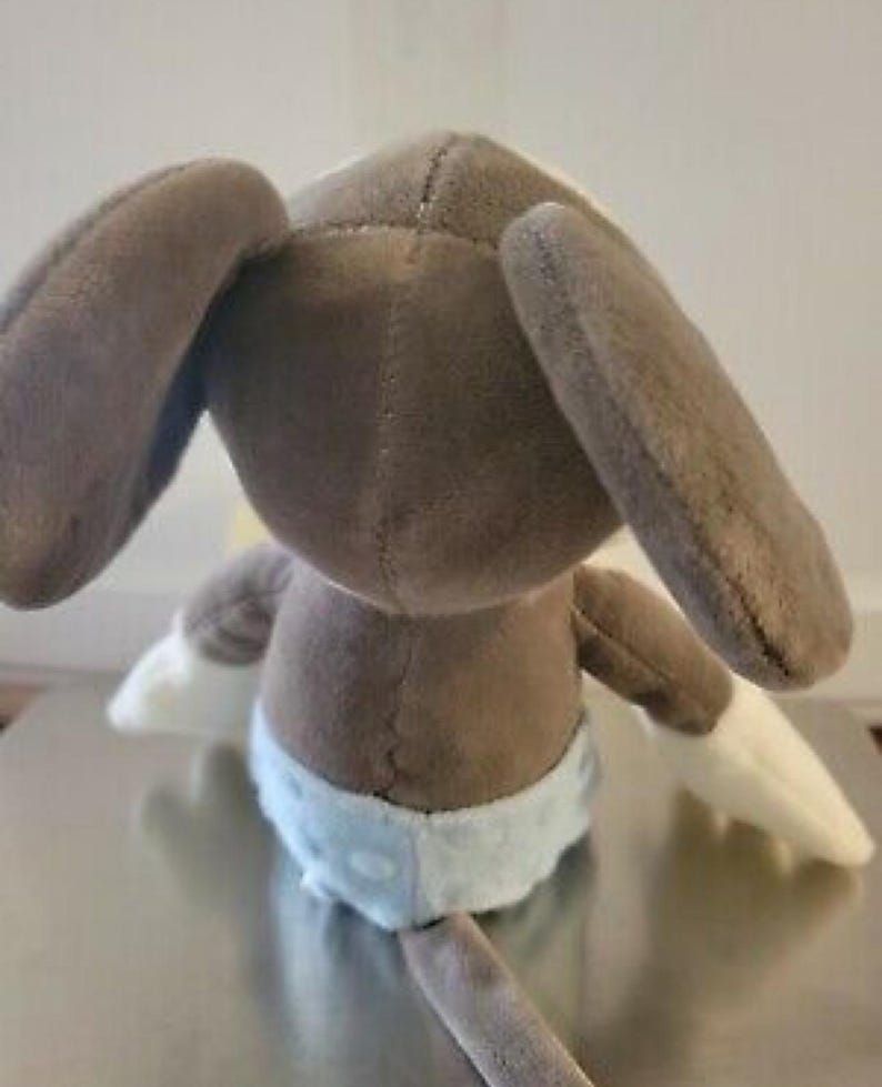 May include: A plush toy bunny, viewed from the back, featuring long, floppy ears and a brown body. The bunny wears a light blue diaper with white trim and has white paws. The toy is made of soft, plush material.