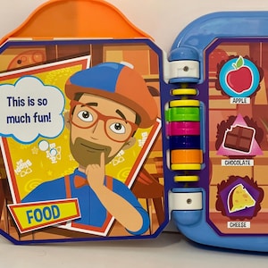 May include: A colorful children's interactive toy book featuring a cartoon character and food illustrations. The book has a blue and orange plastic frame with the text "This is so much fun!" and the word "FOOD". The right side shows pictures of food.