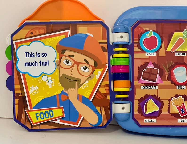 May include: A colorful children's interactive toy book featuring a cartoon character and food illustrations. The book has a blue and orange plastic frame with the text "This is so much fun!" and the word "FOOD". The right side shows pictures of food.