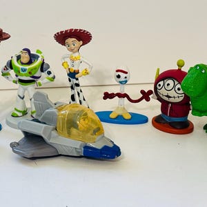Peut inclure: Collection de figurines de personnages de Toy Story, dont Woody, Buzz l'&Eacute;clair, Jessie et Rex le dinosaure. Les figurines sont de couleurs vives et se tiennent sur une surface blanche. Un avion jouet est &eacute;galement inclus.