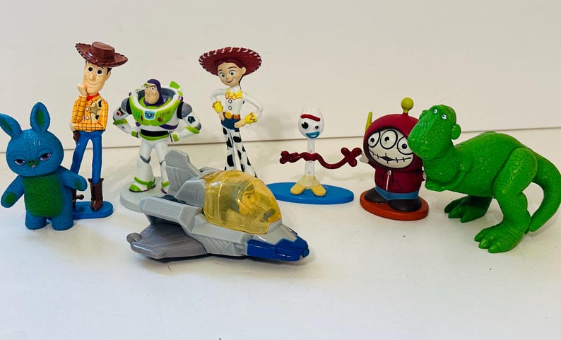Peut inclure: Collection de figurines de personnages de Toy Story, dont Woody, Buzz l'&Eacute;clair, Jessie et Rex le dinosaure. Les figurines sont de couleurs vives et se tiennent sur une surface blanche. Un avion jouet est &eacute;galement inclus.