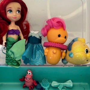 May include: Ariel doll with red hair, a purple top, and a green mermaid tail. Also included are a doll dress, a yellow fish, an orange character with pink hair, a red crab, a hairbrush, and a blue bow. The items are in a clear plastic container.