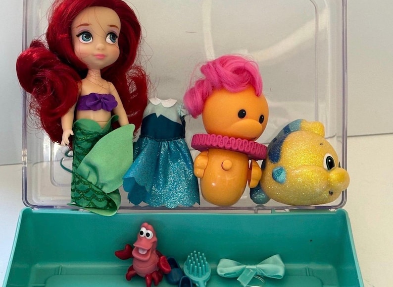 May include: Ariel doll with red hair, a purple top, and a green mermaid tail. Also included are a doll dress, a yellow fish, an orange character with pink hair, a red crab, a hairbrush, and a blue bow. The items are in a clear plastic container.