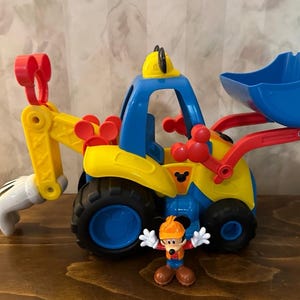 May include: A colorful toy construction vehicle with a blue scoop, yellow body, and red accents. A small Mickey Mouse figure in a construction hat stands in front of the vehicle. The toy has black wheels and a Mickey Mouse logo.