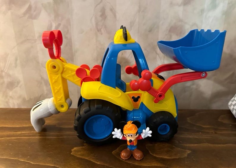 May include: A colorful toy construction vehicle with a blue scoop, yellow body, and red accents. A small Mickey Mouse figure in a construction hat stands in front of the vehicle. The toy has black wheels and a Mickey Mouse logo.