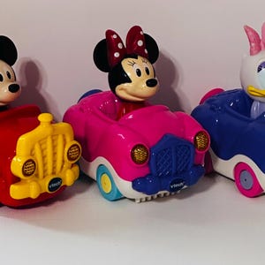 May include: Three VTech toy vehicles featuring Mickey Mouse in a red car, Minnie Mouse in a pink car, and Daisy Duck in a blue car. Each character is molded in plastic and sits in the driver seat of their respective colorful, rounded toy automobile.
