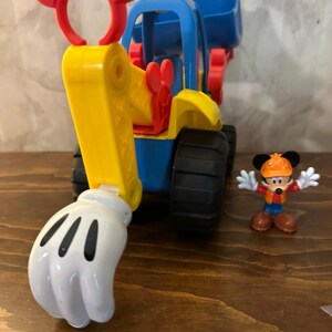May include: A colorful toy construction vehicle with a Mickey Mouse theme. The vehicle is primarily blue and yellow, with red accents and a white glove-shaped scoop. A small Mickey Mouse figurine stands nearby.