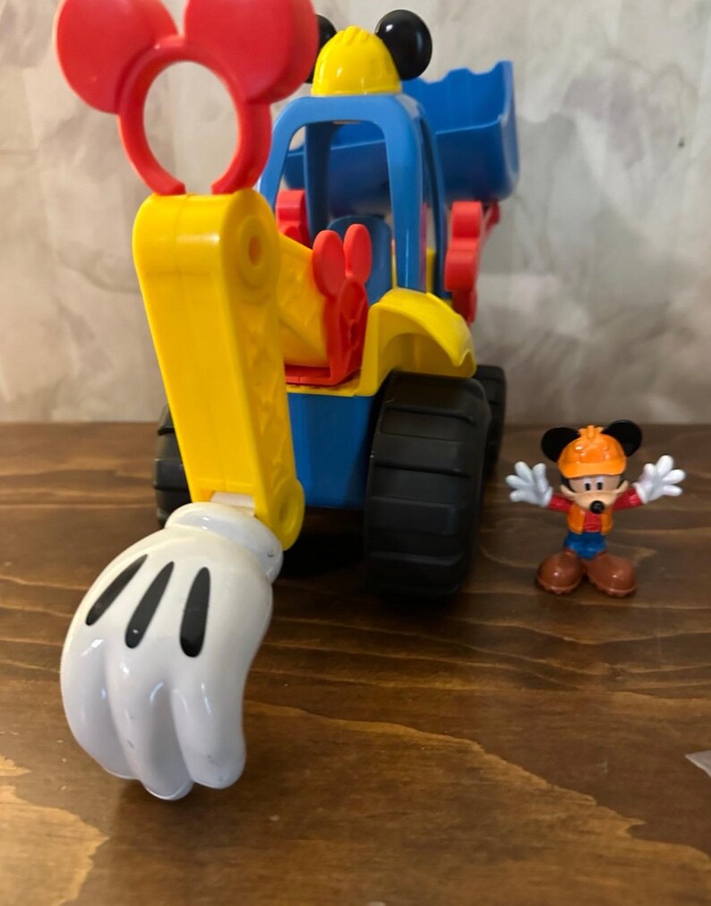 May include: A colorful toy construction vehicle with a Mickey Mouse theme. The vehicle is primarily blue and yellow, with red accents and a white glove-shaped scoop. A small Mickey Mouse figurine stands nearby.