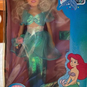 May include: A Disney's The Little Mermaid doll of Arista in a box. The doll is wearing a teal dress with a matching bow. The box features images of Ariel and the text "From Disney's Award-winning Movie!"