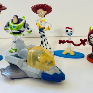 Peut inclure: Collection de figurines de personnages de Toy Story, dont Buzz l'&Eacute;clair, Jessie et Forky. Buzz l'&Eacute;clair est dans un vaisseau spatial. Les figurines sont de couleurs vives et se tiennent sur une surface blanche.