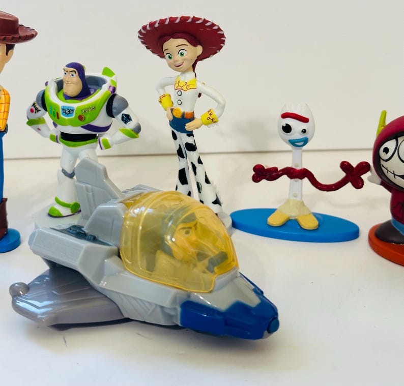 Peut inclure: Collection de figurines de personnages de Toy Story, dont Buzz l'&Eacute;clair, Jessie et Forky. Buzz l'&Eacute;clair est dans un vaisseau spatial. Les figurines sont de couleurs vives et se tiennent sur une surface blanche.