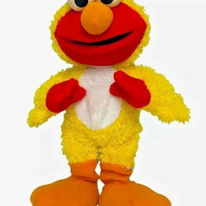 May include: A plush toy in the shape of a yellow chicken with red accents. The toy has large, round eyes, an orange beak, and red hands and feet. The chest is white.