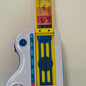 Pode incluir: Uma guitarra de brinquedo colorida com corpo branco e bra&ccedil;o amarelo. O bra&ccedil;o apresenta gr&aacute;ficos de animais e o corpo possui um alto-falante, bot&otilde;es e uma luz vermelha. O brinquedo &eacute; da marca "baby einstein-".