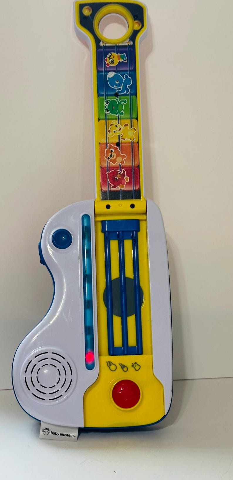 Pode incluir: Uma guitarra de brinquedo colorida com corpo branco e bra&ccedil;o amarelo. O bra&ccedil;o apresenta gr&aacute;ficos de animais e o corpo possui um alto-falante, bot&otilde;es e uma luz vermelha. O brinquedo &eacute; da marca "baby einstein-".