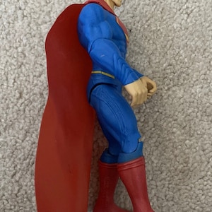 May include: A Superman action figure in a side profile view. The figure is wearing a blue suit with red boots and a red cape. The figure has black hair and a determined expression.