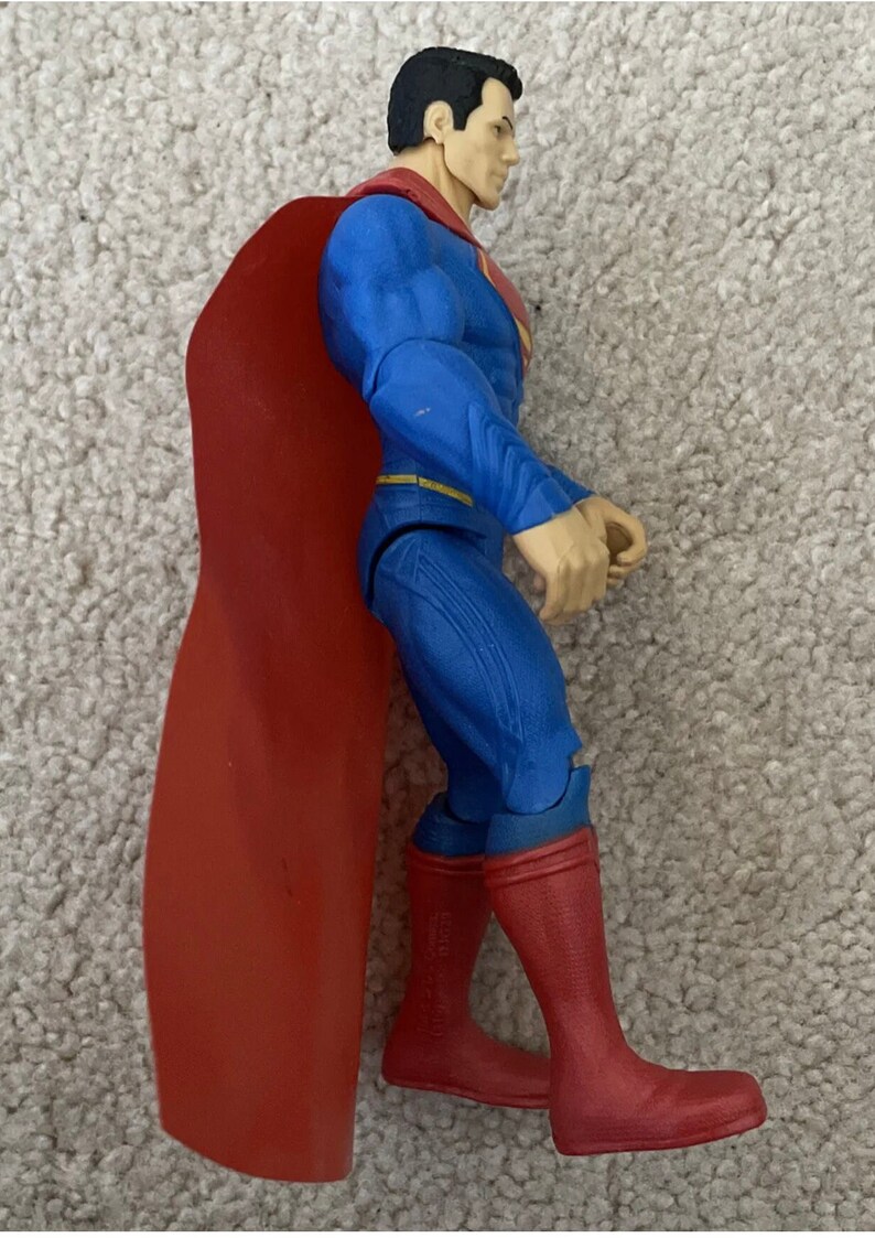 May include: A Superman action figure in a side profile view. The figure is wearing a blue suit with red boots and a red cape. The figure has black hair and a determined expression.