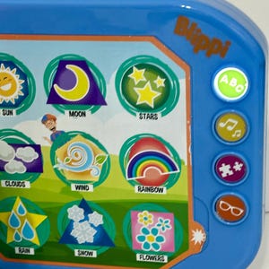 May include: A blue Blippi-branded toy with colorful illustrations of weather elements like sun, moon, stars, clouds, wind, rainbow, rain, snow, and flowers. It has buttons with ABC, music notes, puzzle pieces, and glasses icons.
