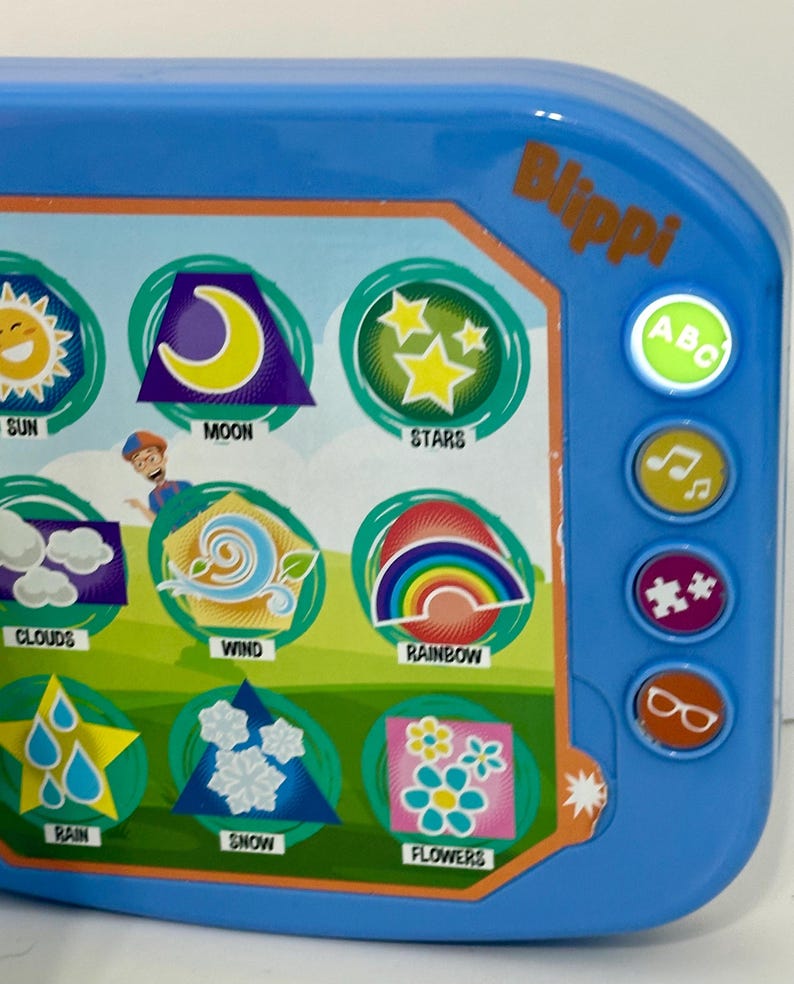 May include: A blue Blippi-branded toy with colorful illustrations of weather elements like sun, moon, stars, clouds, wind, rainbow, rain, snow, and flowers. It has buttons with ABC, music notes, puzzle pieces, and glasses icons.