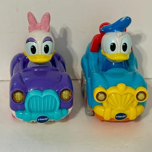 May include: Two toy cars featuring Disney characters Daisy Duck and Donald Duck. Daisy's car is purple with a teal bumper and yellow headlights. Donald's car is blue with a yellow front and headlights. Both cars have the Vtech logo.