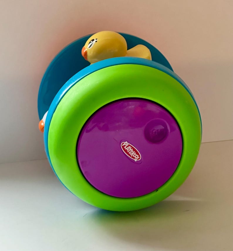 May include: A Playskool toy with a yellow duck on top. The toy is a combination of bright colors, including teal, green, and purple. The Playskool logo is visible on the purple base. The toy is designed for young children.