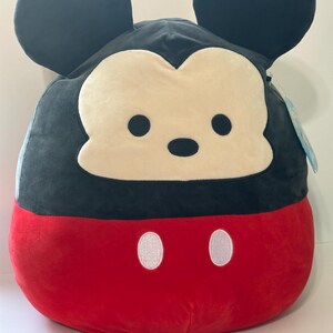 Op de afbeelding: Een pluche Mickey Mouse-kussen. Het kussen heeft een zwarte kop en oren, een cr&egrave;mekleurig gezicht, zwarte ogen en een rode onderkant met twee witte knoopdetails. Het kussen is zacht en rond.