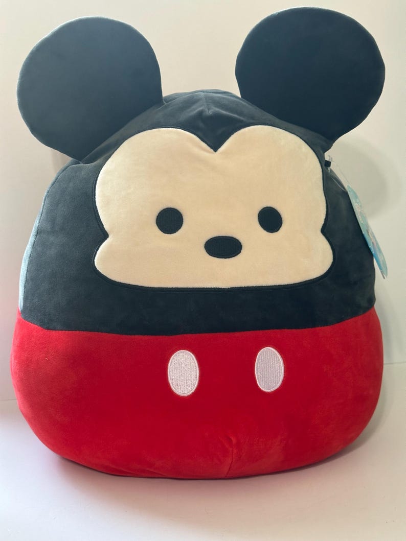 Op de afbeelding: Een pluche Mickey Mouse-kussen. Het kussen heeft een zwarte kop en oren, een cr&egrave;mekleurig gezicht, zwarte ogen en een rode onderkant met twee witte knoopdetails. Het kussen is zacht en rond.