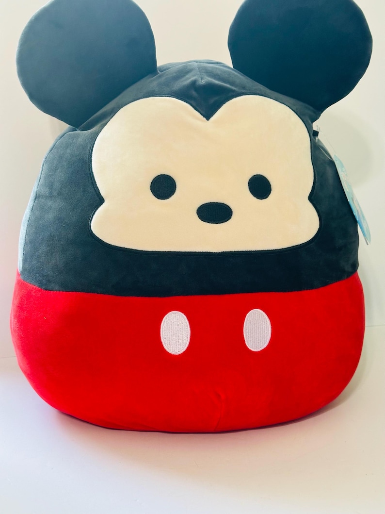 Op de afbeelding: Een pluche Mickey Mouse-kussen. Het kussen heeft een zwarte kop en oren, een cr&egrave;mekleurig gezicht en een rode body met twee witte knoopdetails. Het kussen is zacht en rond.