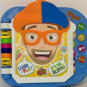 May include: A blue and white children's learning toy featuring a cartoon character with orange glasses and a hat. The toy has buttons with ABCs, music notes, and other symbols. Text on the toy says "Learn & Play" and "It's Me, Blippi!"
