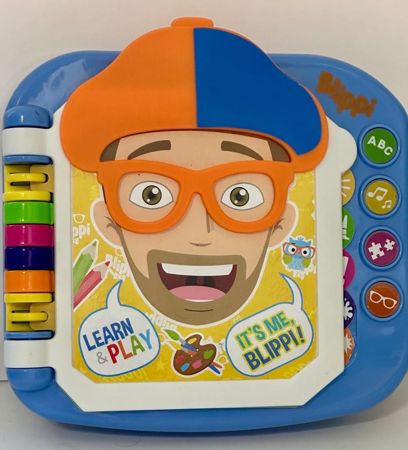 May include: A blue and white children's learning toy featuring a cartoon character with orange glasses and a hat. The toy has buttons with ABCs, music notes, and other symbols. Text on the toy says "Learn & Play" and "It's Me, Blippi!"