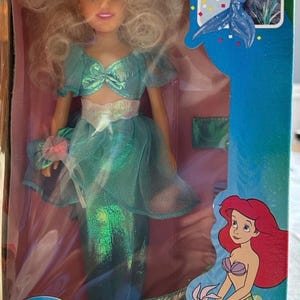 May include: A boxed Disney's The Little Mermaid doll of Ariel's sister, Arista. The doll wears a teal mermaid outfit with a matching bow. The box features the movie's logo and a picture of Ariel.