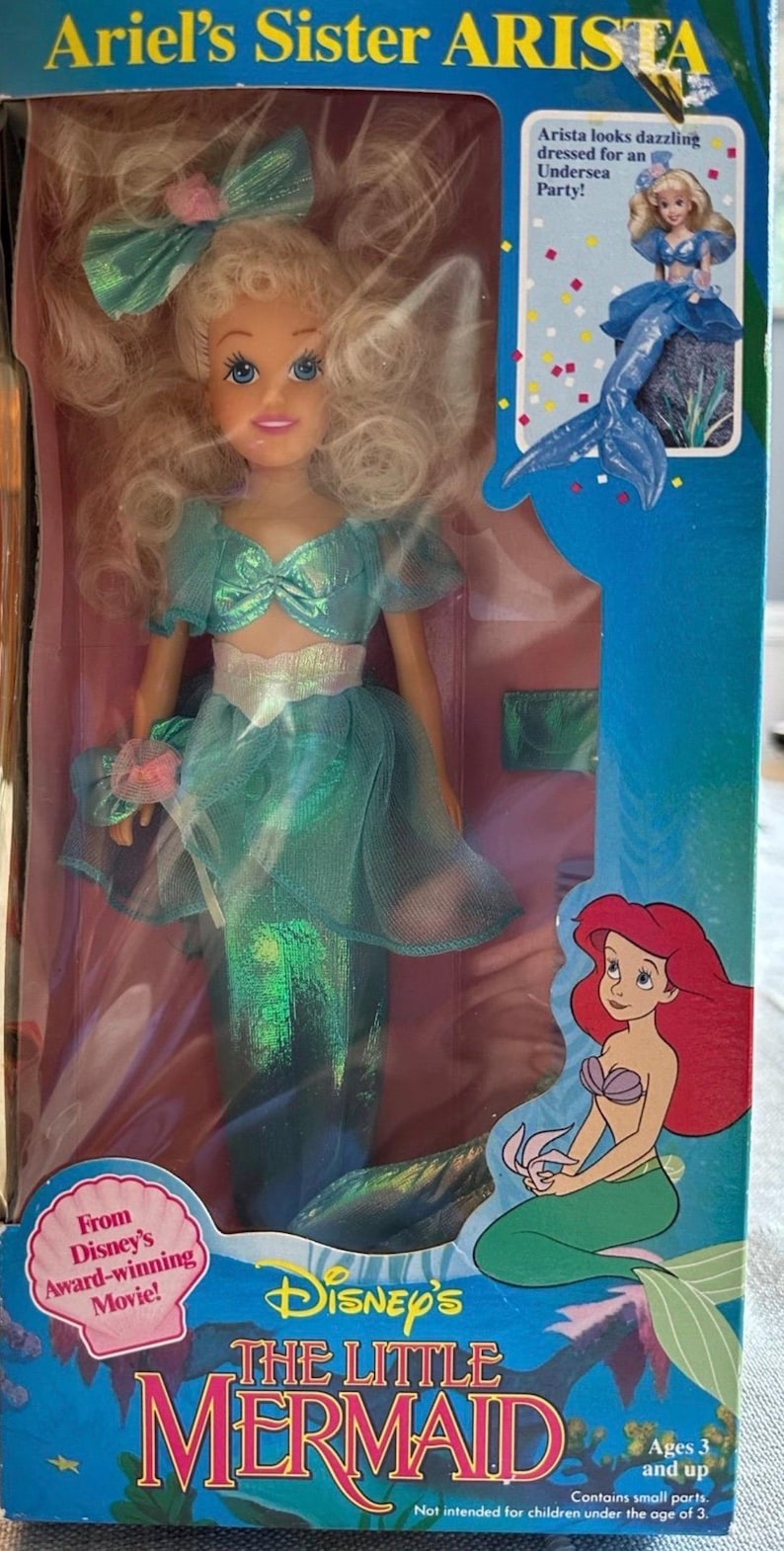 May include: A boxed Disney's The Little Mermaid doll of Ariel's sister, Arista. The doll wears a teal mermaid outfit with a matching bow. The box features the movie's logo and a picture of Ariel.